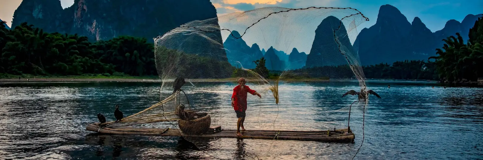 A cormorant fisherman casting his net from a bamboo raft on the Li River near Yangshuo, with karst limestone peaks rising behind