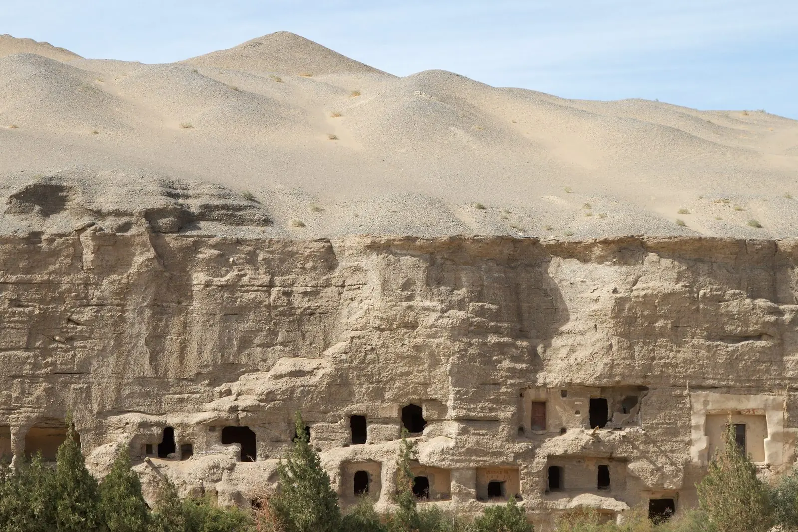 Rows of cave openings cut into the cliff face of the Mogao Grottoes near Dunhuang in Gansu, desert hills rising behind the cliff