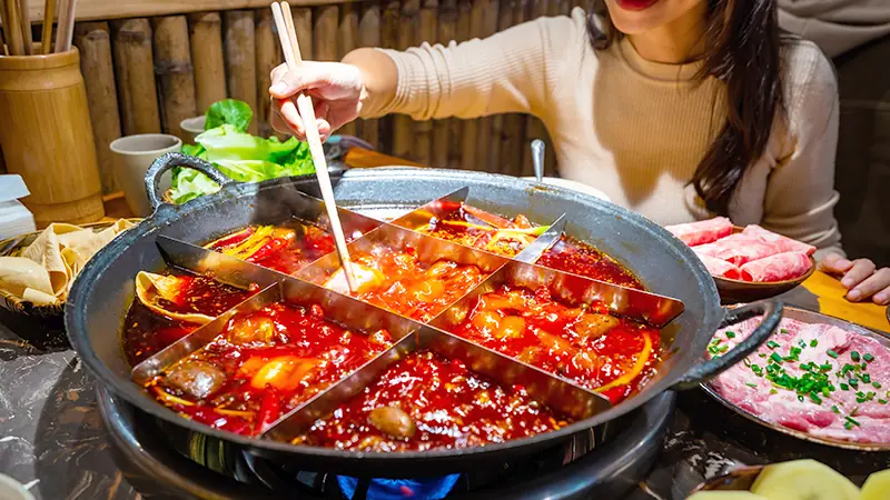 A Chongqing nine-grid Sichuan hotpot — a diner lifting food from the chilli broth with chopsticks, sliced meat and greens alongside