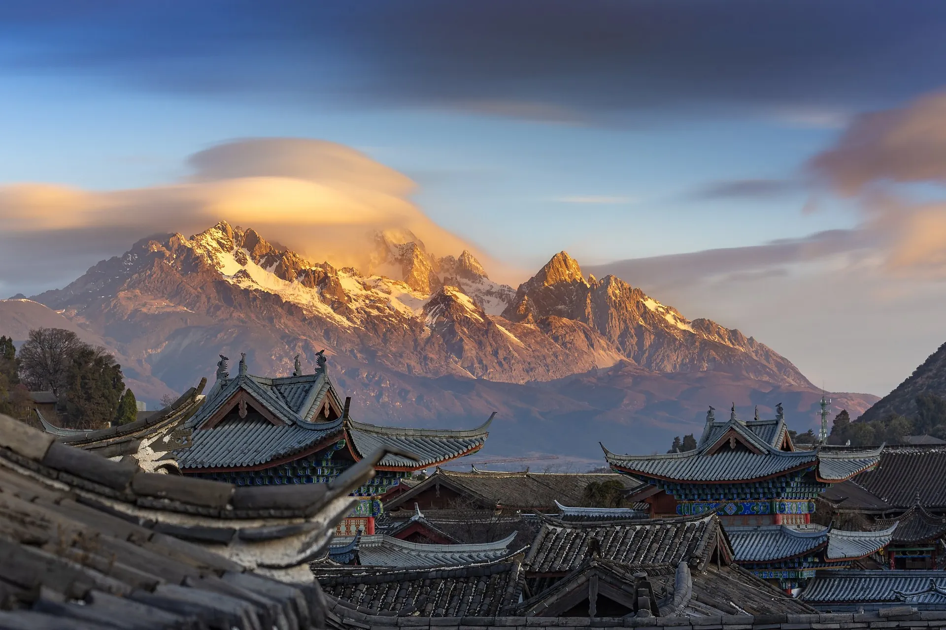 Lijiang's tiled rooftops in golden dawn light beneath the snow peaks of Jade Dragon Snow Mountain in Yunnan